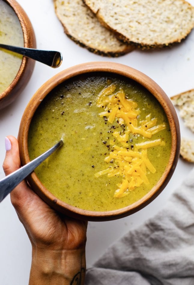 A hand holding a wooden bowl of broccoli and cheese soup with bread slices in the background.