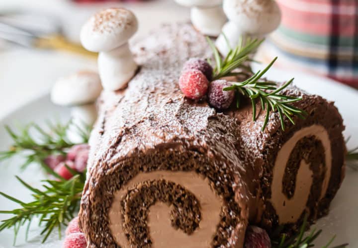 Yule log cake garnished with greens & berries, on a white platter.