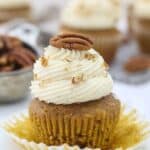 A pumpkin cupcake with the wrapper pulled down, it's frosted with a pretty swirled frosting and topped with a pecan.