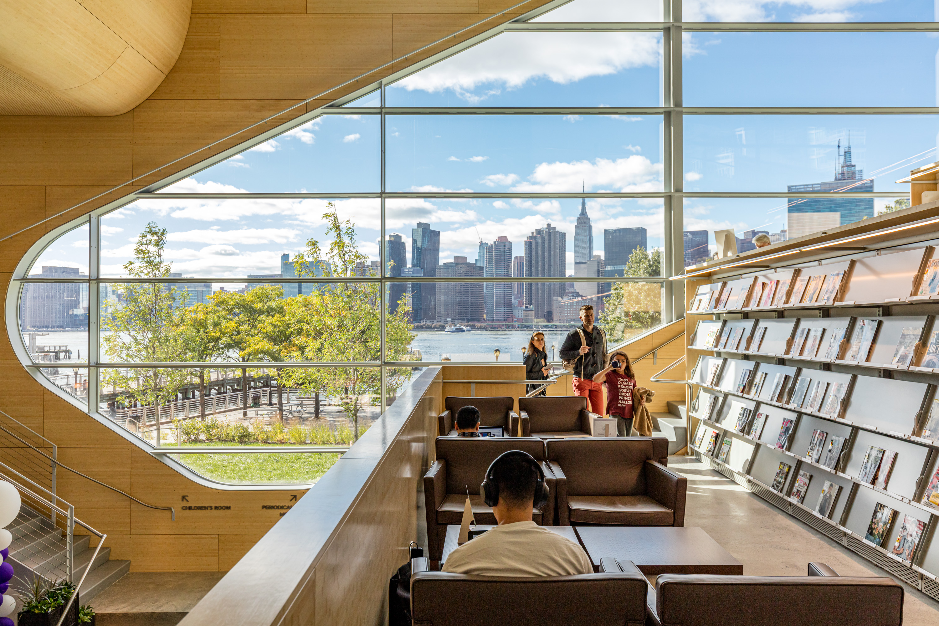 View of Manhattan&rsquo;s skyline out of a curved window at the Hunters Point library. The wall is a light wood, there are stacks filled with books, and seats with tables for studying.