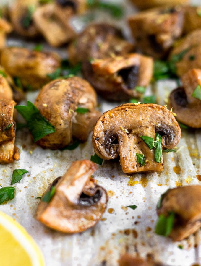 Close up of a balsamic roasted mushrooms on a baking sheet surrounded by more mushrooms. They are topped with fresh parsley.