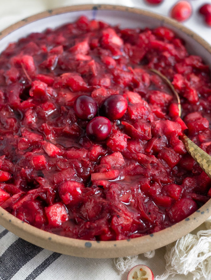 Bowl of homemade cranberry sauce topped with 3 whole cranberries and spoon coming out of it. It is on a towel with cranberries behind it.