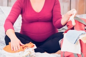 A pregnant person wearing a red top and black pants is sitting on a bed, packing baby clothes into their hospital bag. They are sorting through a stack of small garments and holding a piece of baby clothing in one hand, meticulously following the hospital bag checklist.