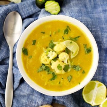 A bowl of curried cauliflower soup on a blue towel with a spoon, garnished with cauliflower florets and grated zucchini.
