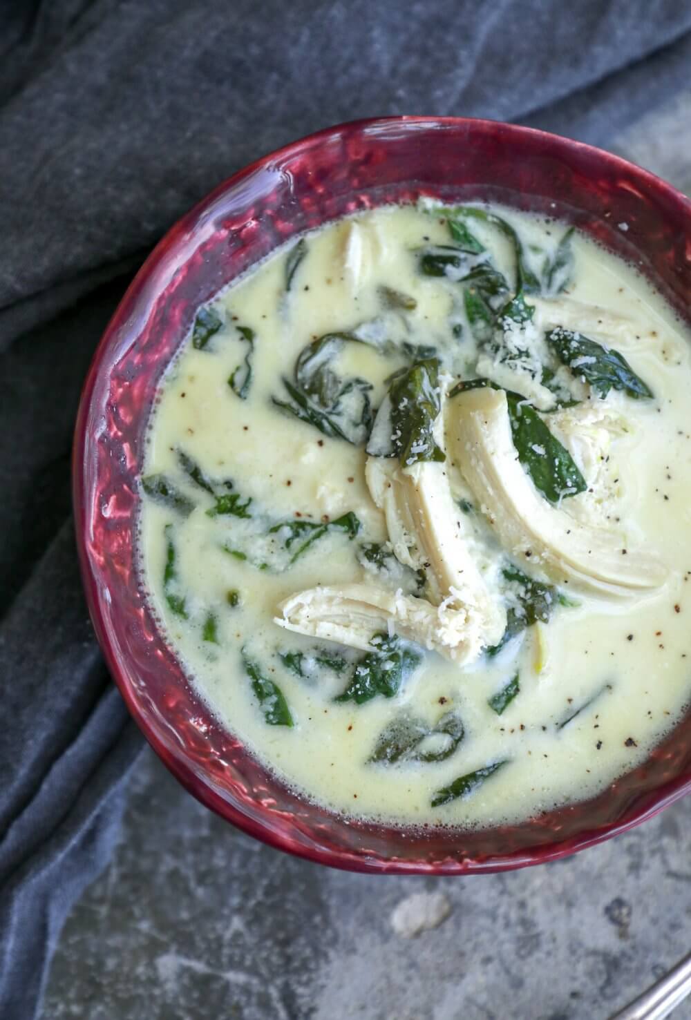 keto chicken Florentine soup in a red bowl on a grey concrete background with a grey napkin shot from above with chunks of chicken on top