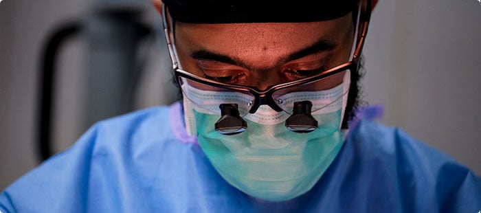 A surgeon in a mask and glasses with loupes performs surgery